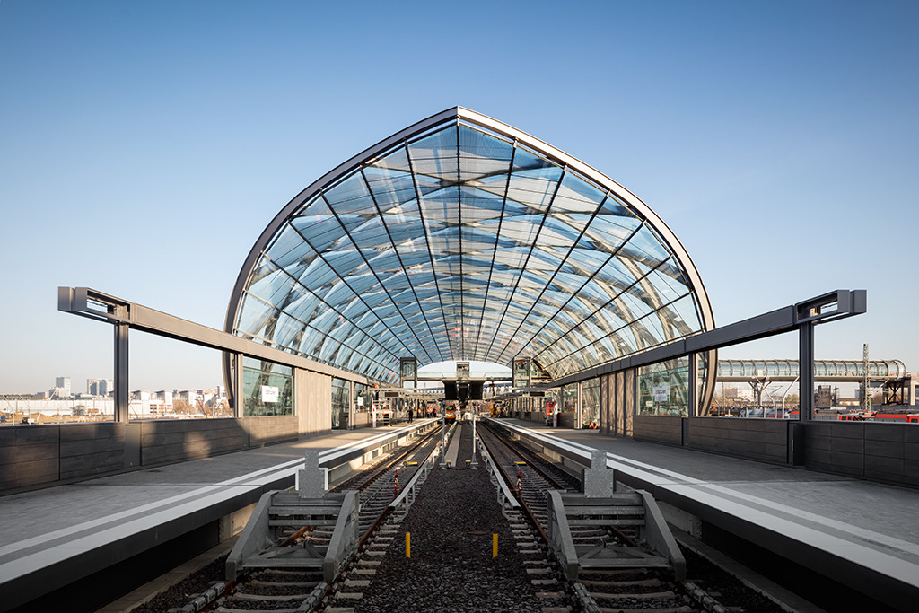Empgangsgeste für die einfahrenden Bahnen (Bild: Marcus Bredt) Realisierungswettbewerb, Einladungswettbewerb 2012 - 1. Preis (2013) U-Bahnhaltestelle der U4 an den Elbbrücken mit Anbindung an die S-Bahnhaltestelle. Das Quartier „Elbbrücken“ wird den östlichen Abschluss der Hafencity bilden. Um dieses auch effektiv an den öffentlichen Nahverkehr anzuschließen, wird die U-Bahnlinie U4 bis zu den Elbbrücken verlängert. Die tonnenförmige Dachkonstruktion der dort geplanten Haltestelle wird durch gekreuzte, bogenförmige Rahmen gebildet. Die hierdurch entstehende optische Präsenz des Tragwerks wird durch die nach innen abgehängte Verglasung unterstrichen. Die kreuzförmige Anordnung der Rahmen ergeben ein rostartiges System, wodurch eine gegenseitige Stabilisierung der stählernen Bögen gegeben ist. Die lichtdurchflutete Haltestelle ermöglicht so diverse Ausblicke zu den markanten Hochhäusern und zu den Elbbrücken. Entwurf: Volkwin Marg und Jürgen Hillmer mit Stephanie Joebsch Partner: Jürgen Hillmer Projektleitung: Stephanie Joebsch Tragwerksplanung: schlaich bergermann partner Bauherr: Hamburger Hochbahn AG